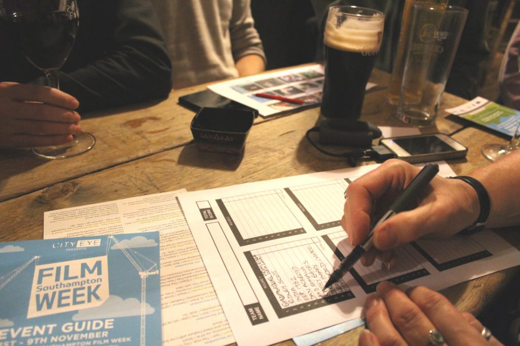 hand writing with pen on quiz sheet. Pub table, low lighting, pint of guinness in shot and another hand holding the stem of a wine glass