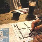hand writing with pen on quiz sheet. Pub table, low lighting, pint of guinness in shot and another hand holding the stem of a wine glass