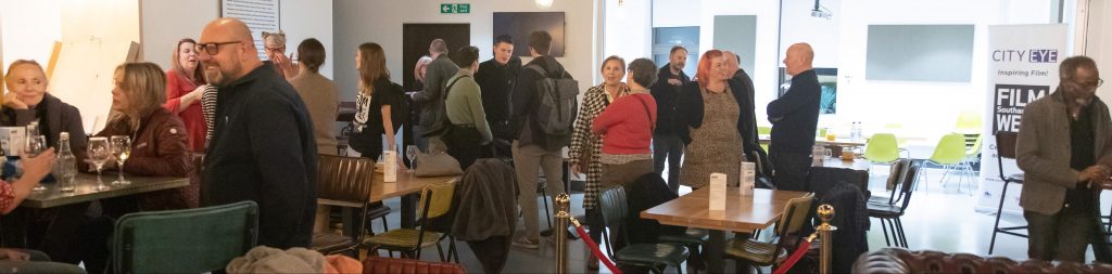 people in a bar standing and sitting, drinking and talking. Can see black and white banner in the top corner that says City Eye and can see red rope splitting part of the room